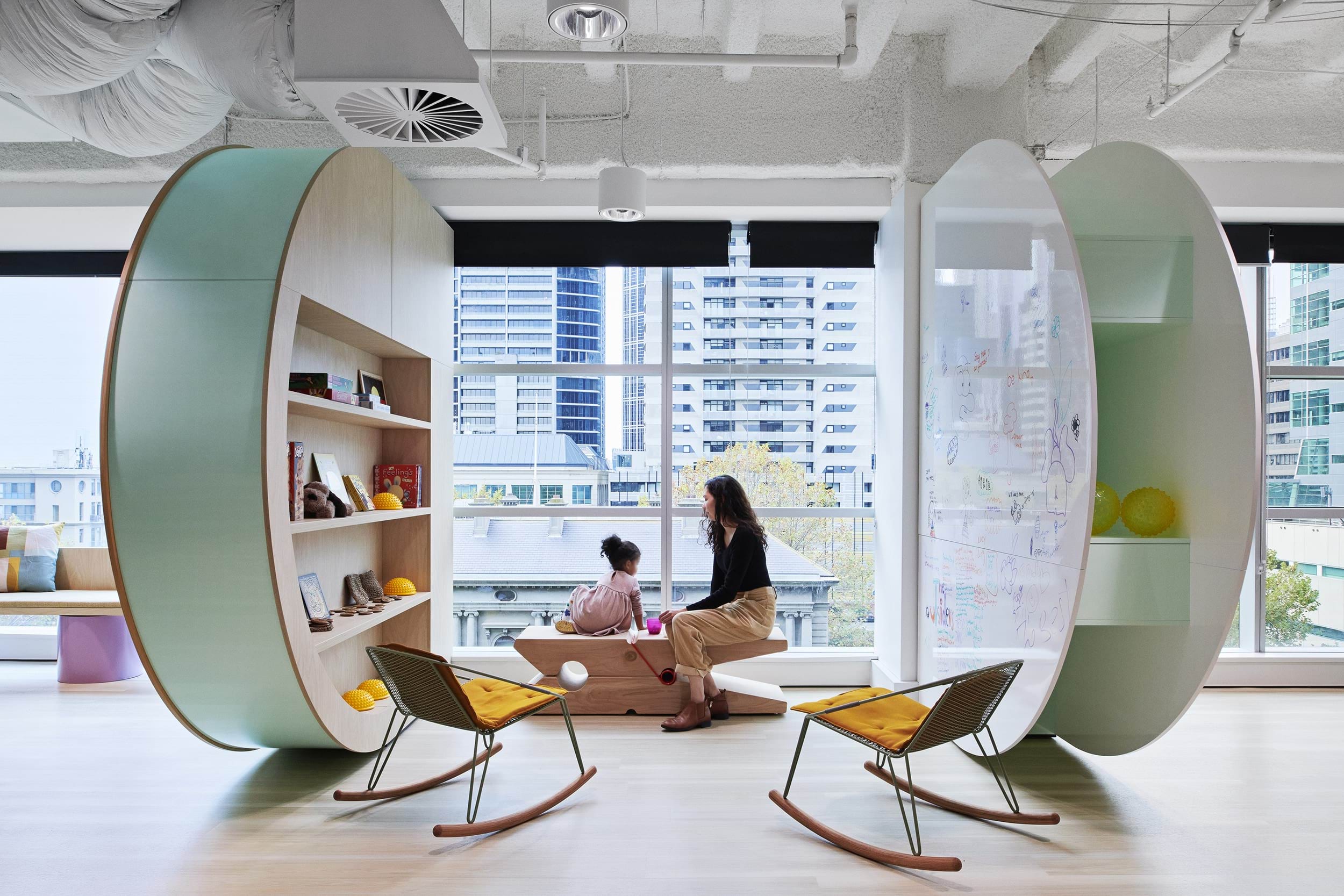 a woman and small child sitting on a bench in a modern building, in front of a wall of windows looking out on a city view. There is a rounded bookshelf on one side of them, and a rounded whiteboard on the other. Two modern rocking chairs complete the area.