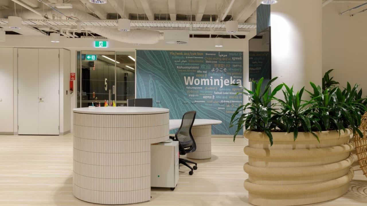 Modern, round reception desk, with exposed ceiling, greenery behind the desk, and a mural of welcoming words in multiple languages on the wall behind.